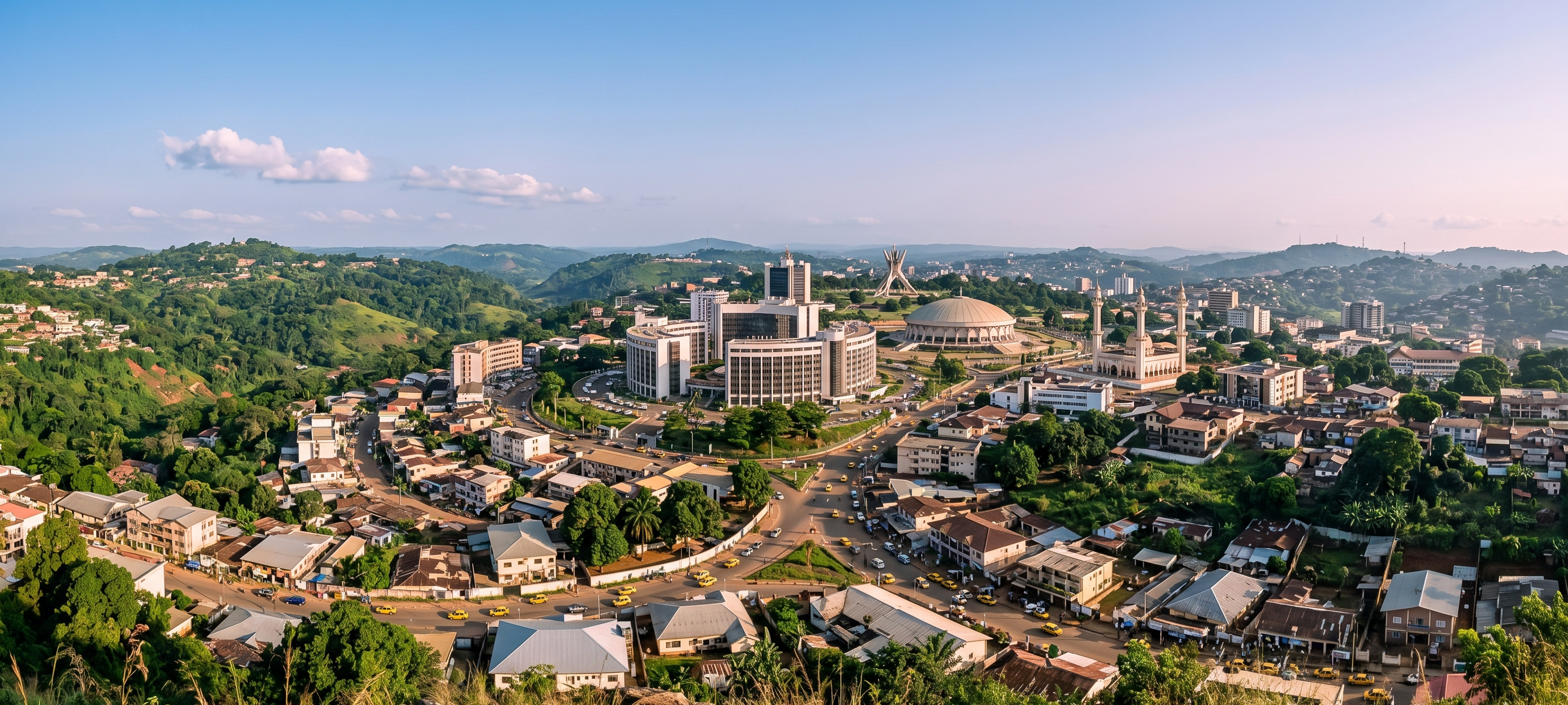 A wide panoramic morning photograph capturing the lush green hills, modern administrative buildings, and landmarks of the Yaoundé, Cameroon, skyline under a clear sky.