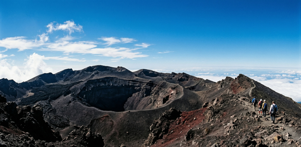 A majestic, eye-level photograph capturing the rugged, barren volcanic craters near the summit of Mount Cameroon, contrasting sharply against a deep blue sky with hikers on the distant rim for scale.
